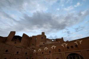 Historic Persian adobe fortress with tall crenellated towers and earthen walls beneath a dramatic cloudy blue sky.