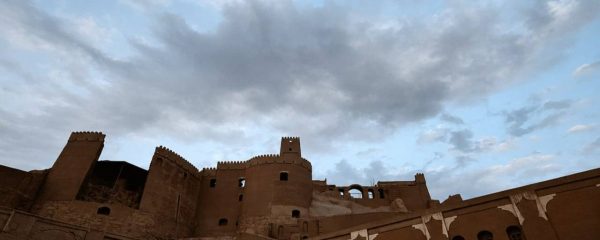 Historic Persian adobe fortress with tall crenellated towers and earthen walls beneath a dramatic cloudy blue sky.